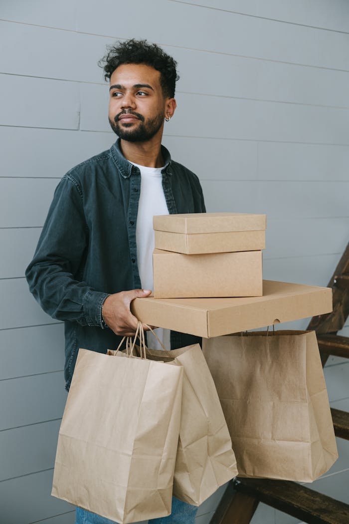 A man holding cardboard boxes and paper bags, looking thoughtful indoors.